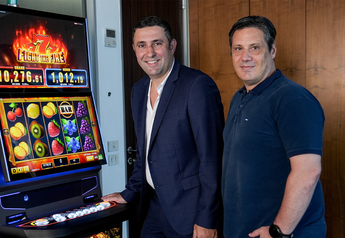 This image shows Pepe Costa and Alexandre Studart posing next to an FBM casino cabinet displaying the slots game Fight the Fire from the FBM Spin & Win collection.
