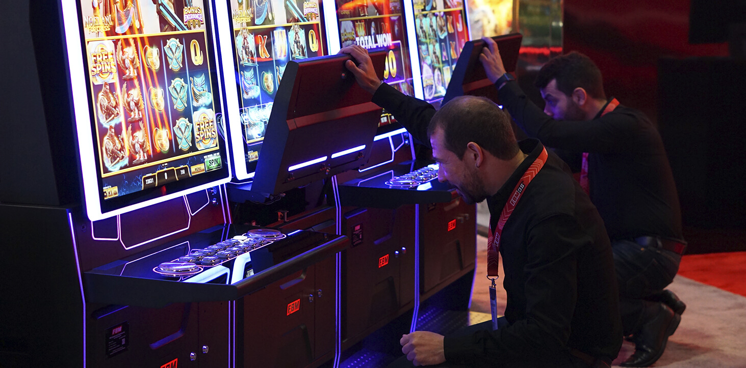 Esta imagen muestra a dos técnicos de FBM realizando acciones de mantenimiento en el gabinete de casino Auria durante una feria comercial.