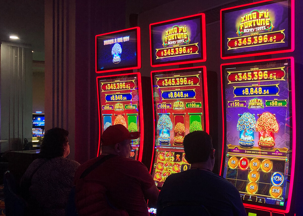 The image shows three Mexican players enjoying FBM's Xing Fú Fortune Money Trees slots in a Mexican casino room.